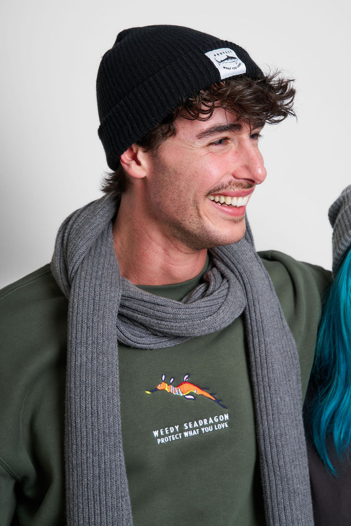 Male model wearing a Kelp relax jumper with a weedy seadragon design and the Protect What You Love text and a lack beanie with a tiger shark design